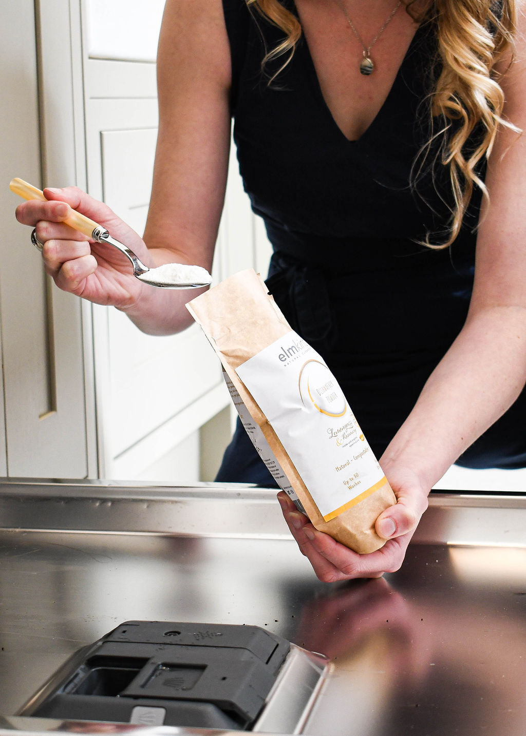 elmkind dishwasher powder 500g, with lemongrass and rosemary essential oils in a brown plastic free paper bag with a woman scooping some powder from the pouch into a dishwasher