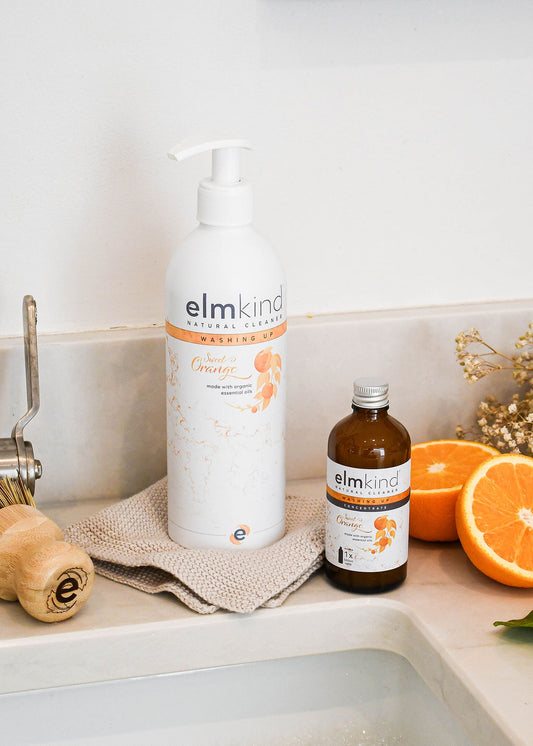 elmkind sweet orange washing up liquid aluminium bottle with white pump on a kitchen sink with a concentrate amber glass refill bottle and some freshly cut oranges