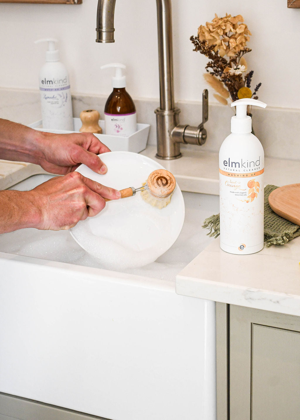 elmkind sweet orange washing up liquid aluminium bottle with white pump on a kitchen sink with a mans hands scrubbing a plate with an elmkind long handled dish brush and lots of bubbles