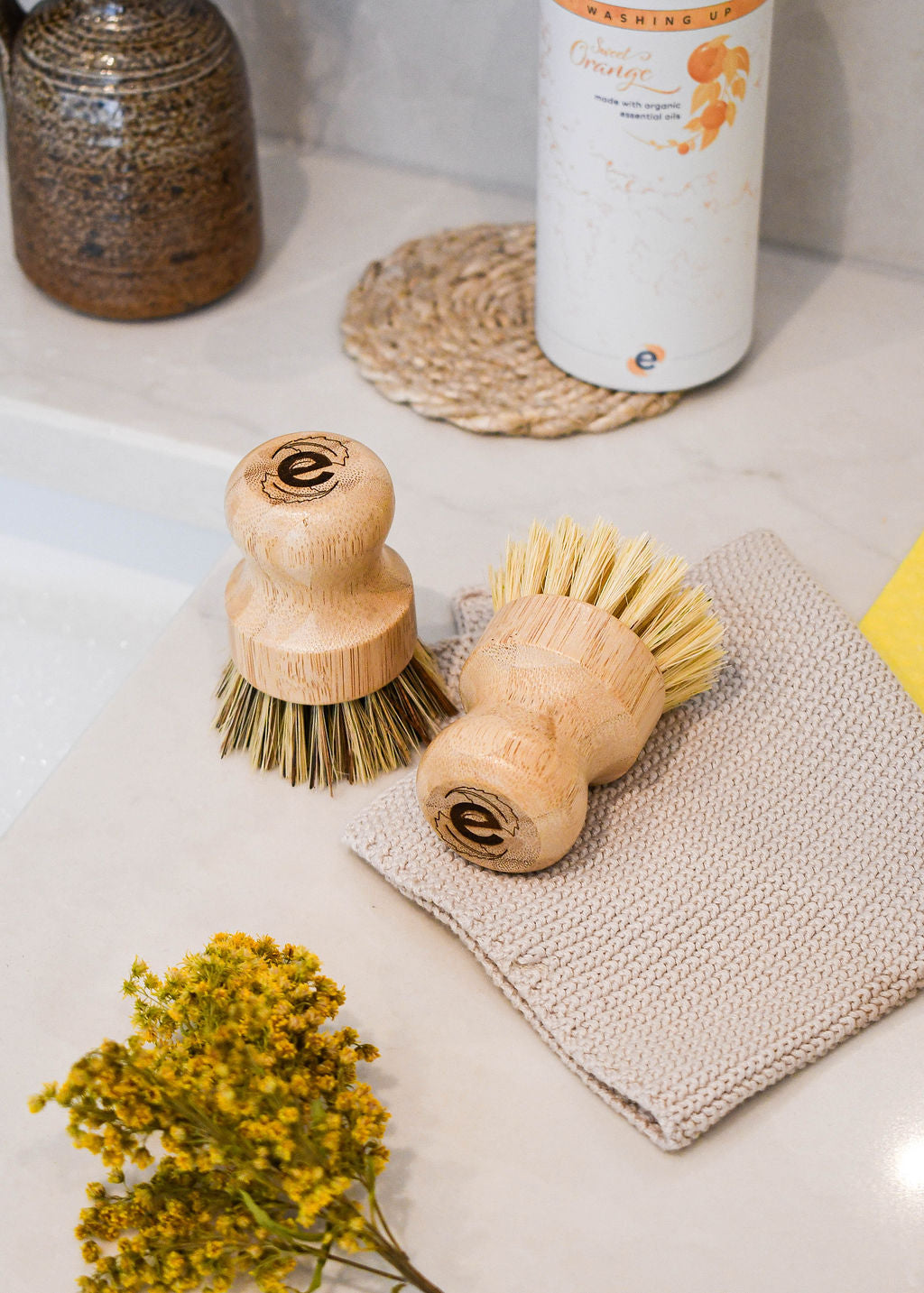 2 elmkind scrubbing brushes, one with soft bristles and one with hard bristles on a kitchen side with some dried flowers and an organic cleaning cloth