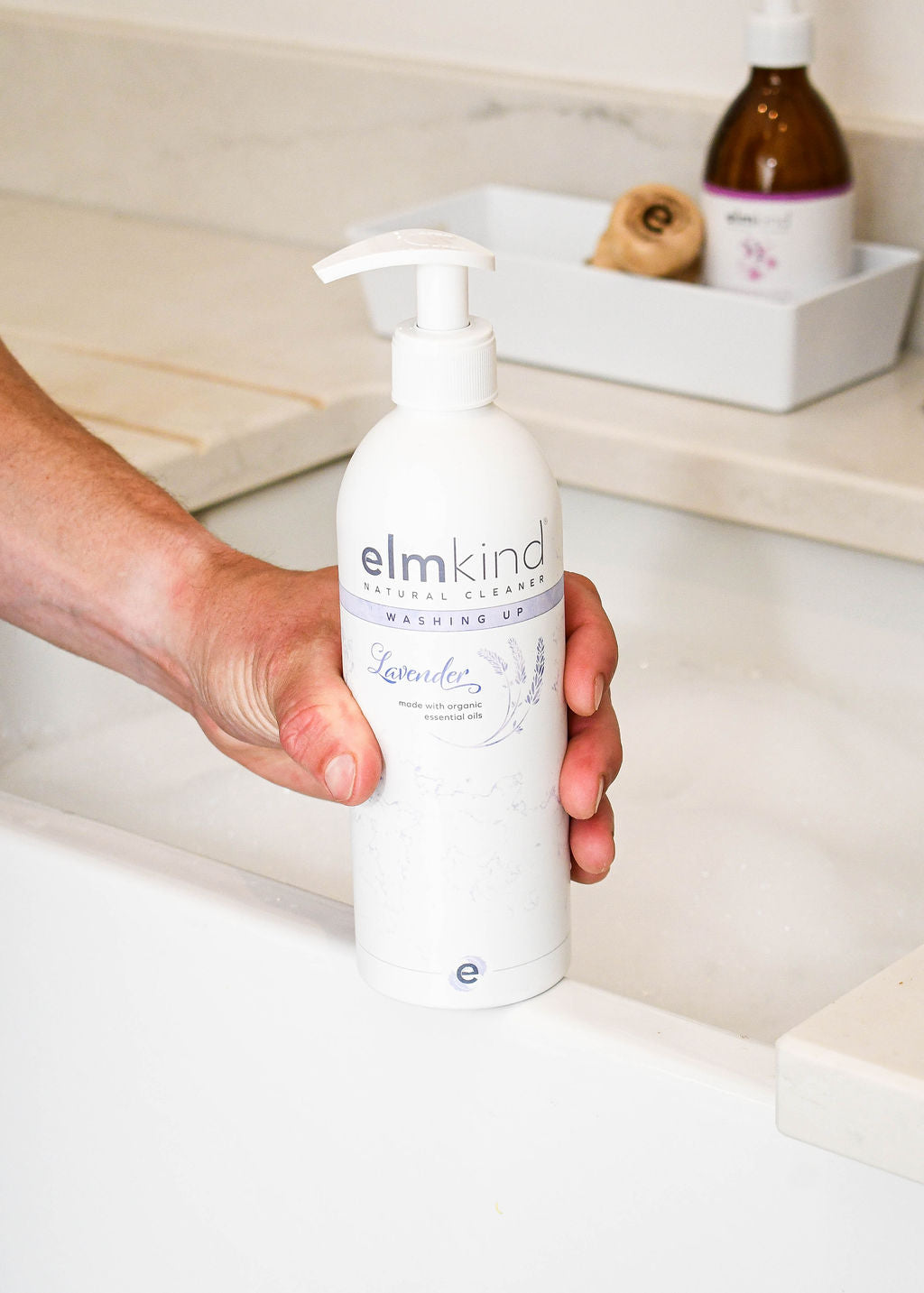 elmkind Lavender washing up liquid with plastic pump resting on a Belfast sink filled with bubbles and with a man holding the bottle