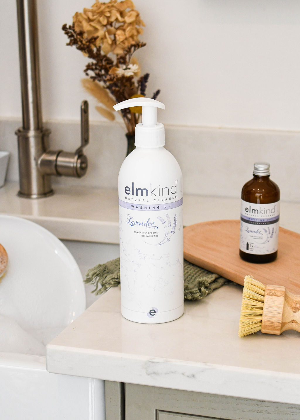 elmkind Lavender washing up liquid with plastic pump on kitchen side with dried flowers, an elmkind washing up liquid refill and an elmkind scrubbing brush next to the sink