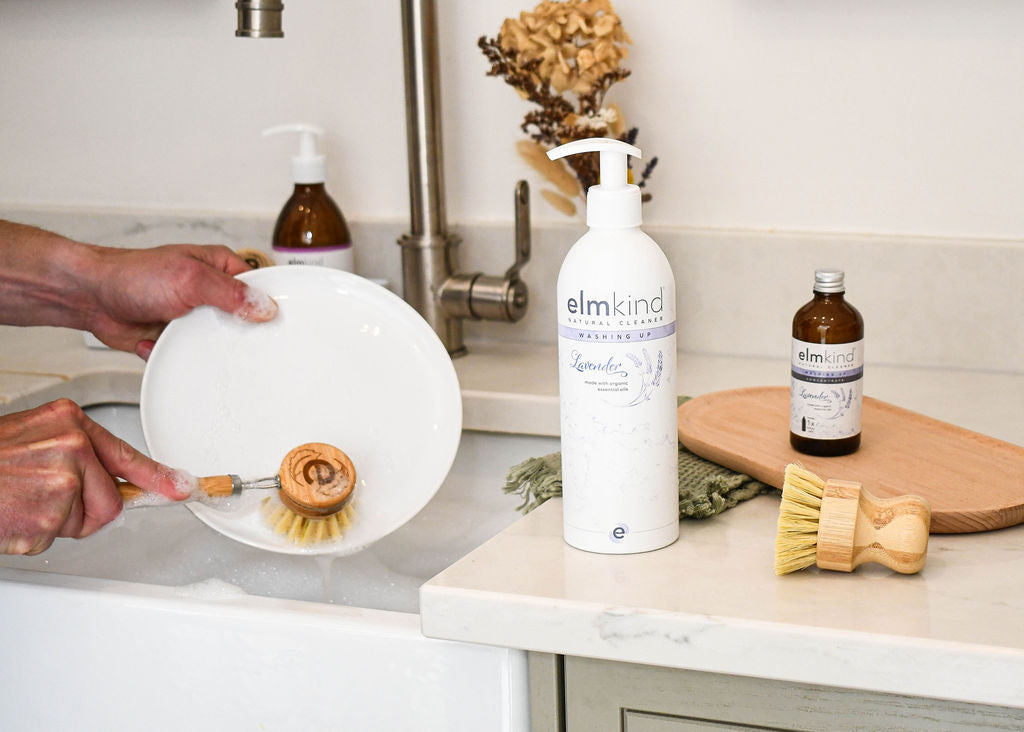 an elmkind lavender washing up liquid in a white aluminium bottle with pump on a kitchen surface with a amber glass refill and a man scrubbing a soap plate with an elmkind long handlebamboo dish brush