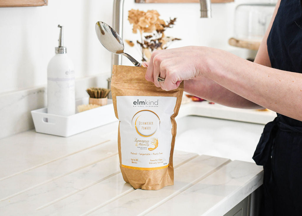 elmkind dishwasher powder 500g, with lemongrass and rosemary essential oils in a brown plastic free paper bag with a woman scooping some powder from the pouch on a kitchen surface
