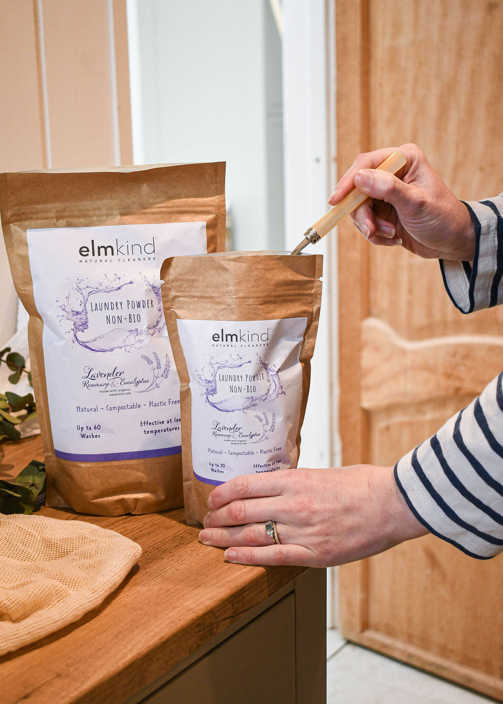 two bags of elmkind laundry powder 1KG and 500g, with lavender, rosemary and eucalyptus essential oils in a brown plastic free paper bag with a woman scooping some powder from the smaller pouch by a washing machine
