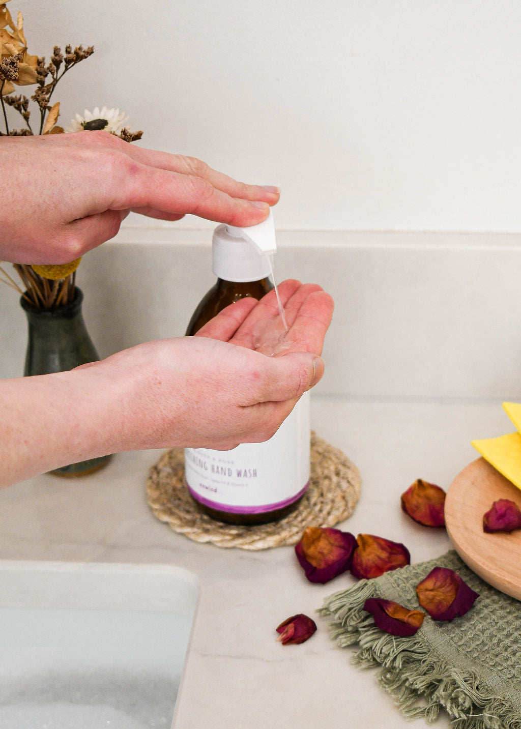 elmkind Lavender and Rose hand wash in a 250ml amber glass bottle with plastic pump squirting into a woman's hand by the sink with some rose petals