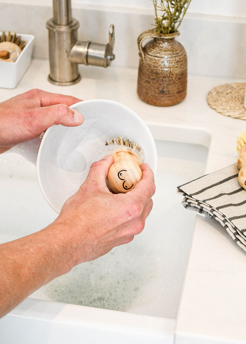 an elmkind scrubbing brush with breasts designed onto the handle to support breast cancer awareness, in a woman's hand scrubbing a soapy plate in a sink
