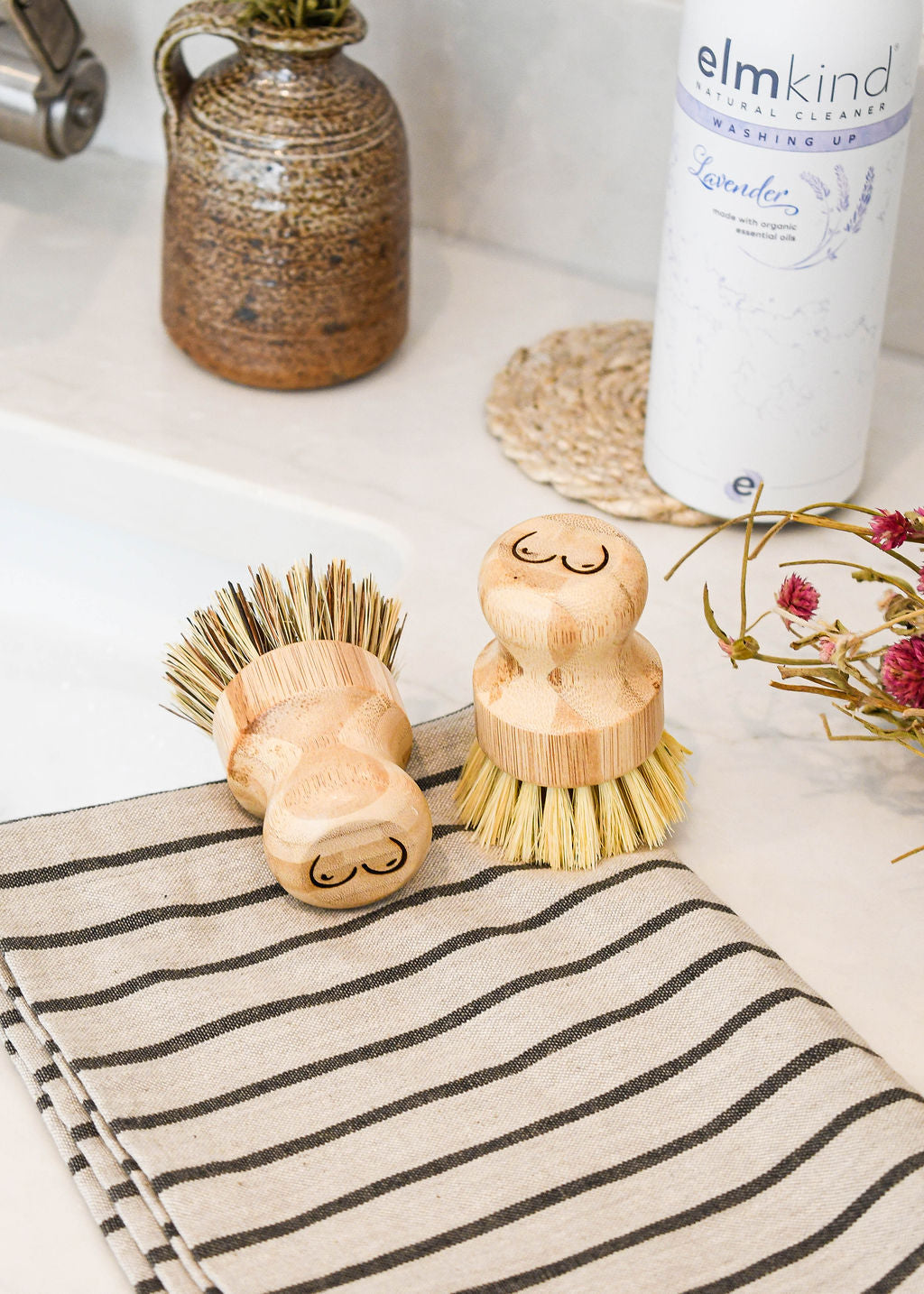 a pair of elmkind scrubbing brushes with breasts designed onto the handle to support breast cancer awareness, sat on a kitchen side with a stripy tea towel