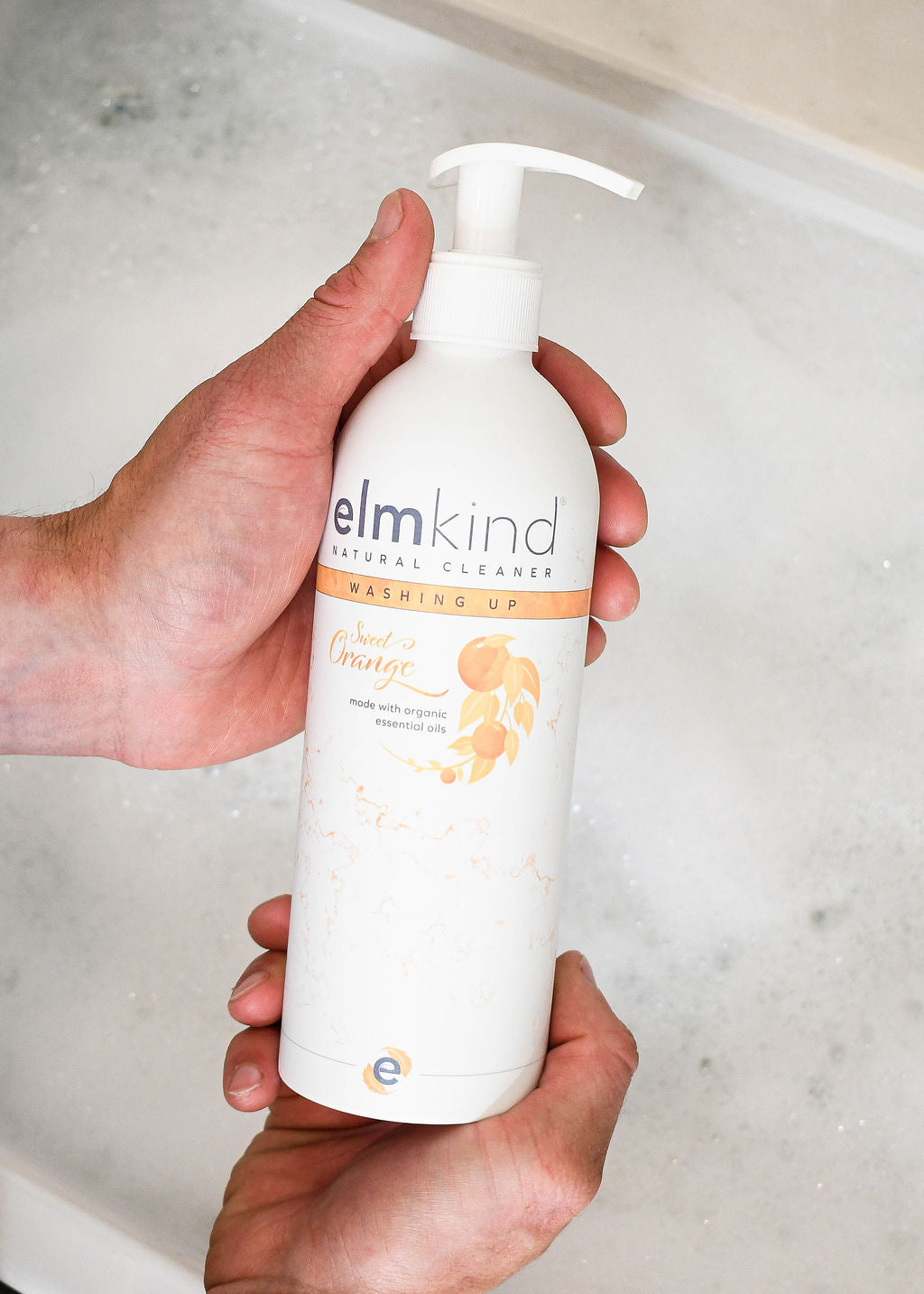 elmkind sweet orange washing up liquid aluminium bottle with white pump in a mans hands over a bubbly sink of powerful natural washing up liquid