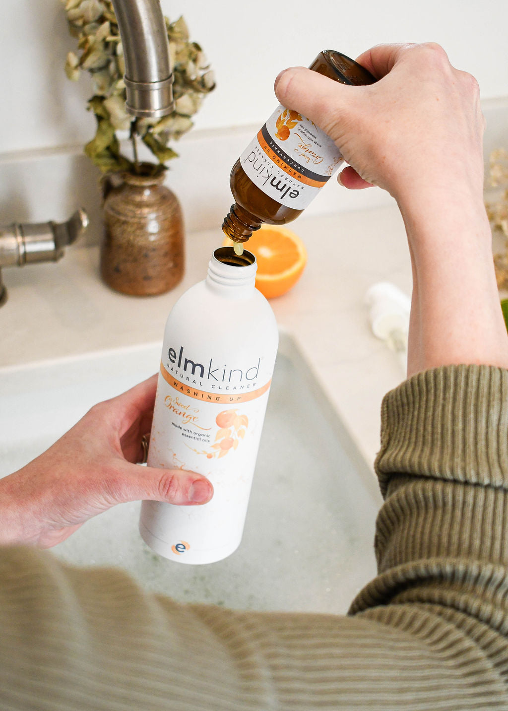 a womans hands holding an elmkind sweet orange washing up liquid aluminium bottle and refilling it with a  plastic free concentrated amber glass refill bottle