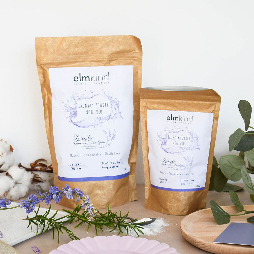 Two bags of elmkind laundry powder 1KG and 500g, with lavender, rosemary and eucalyptus essential oils in a brown plastic free paper bag on a kitchen side with e cotton balls, lavender flowers and rosemary