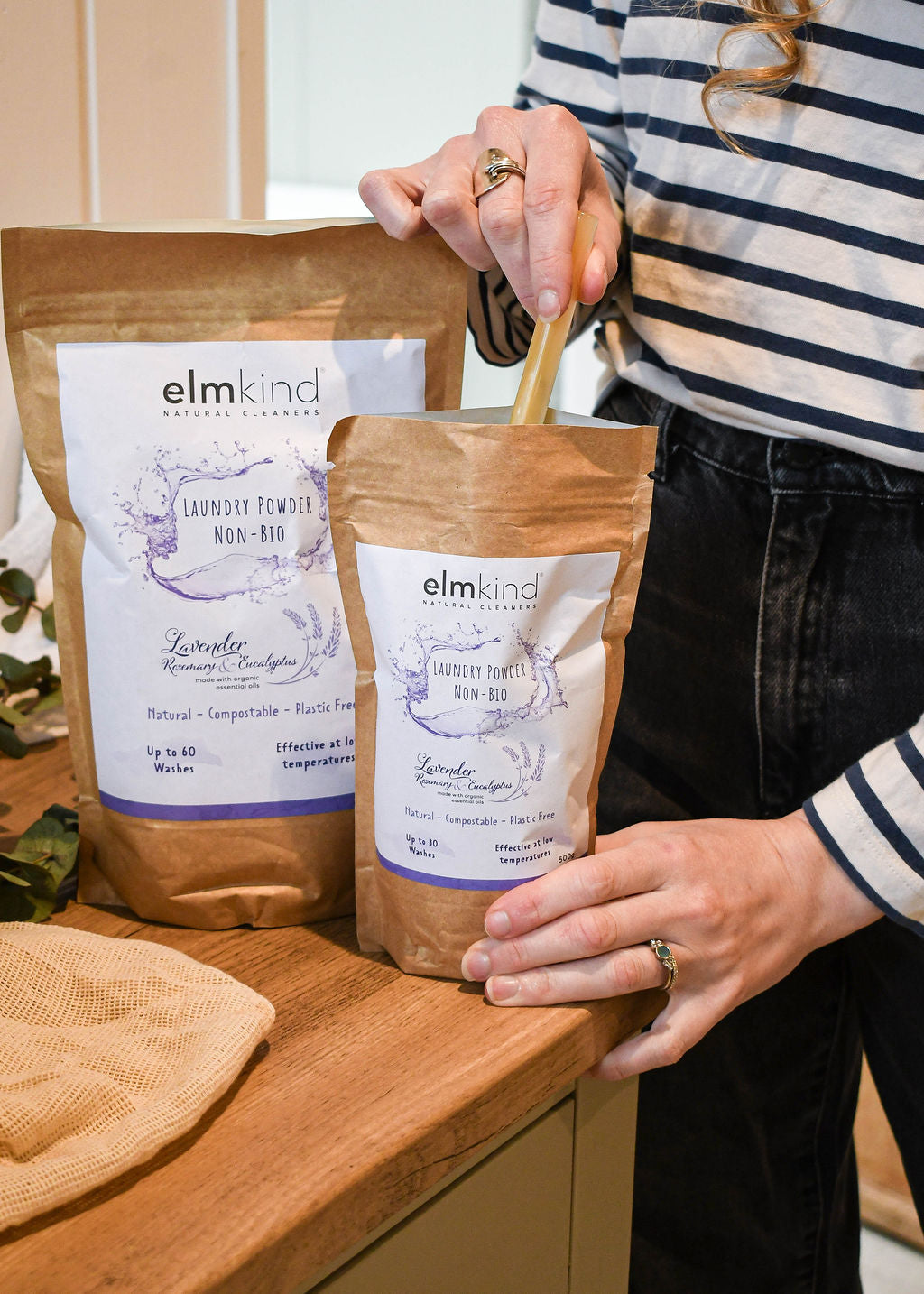 two bags of elmkind laundry powder 1KG and 500g, with lavender, rosemary and eucalyptus essential oils in a brown plastic free paper bag with a woman scooping some powder from the smaller pouch by a washing machine